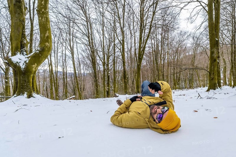 Man wearing a yellow winter jacket and hat, sunglasses, lying relaxed on the snow covered ground in a winter forest, enjoying a peaceful outdoor moment and cold weather