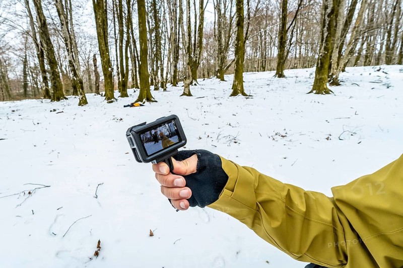 Person holding an action camera showing live footage while hiking through a snow covered forest, capturing pov video of winter wilderness, cold weather gear and frosted trees
