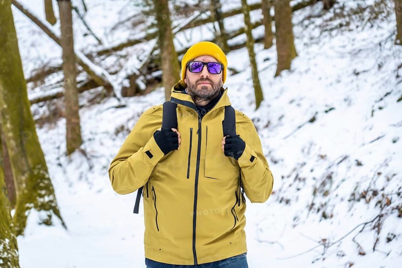 Man in yellow jacket and beanie with sunglasses and backpack hiking through a snow covered forest in winter, enjoying a peaceful, frosty outdoor adventure and scenic landscape