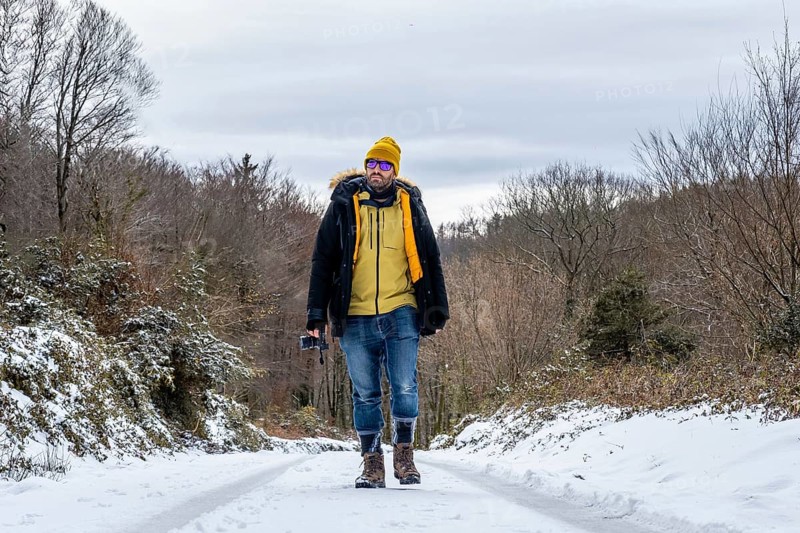 Man in warm winter attire and sunglasses walking on a snowy road through a woodland landscape, holding a compact camera and enjoying outdoor adventure in cold weather
