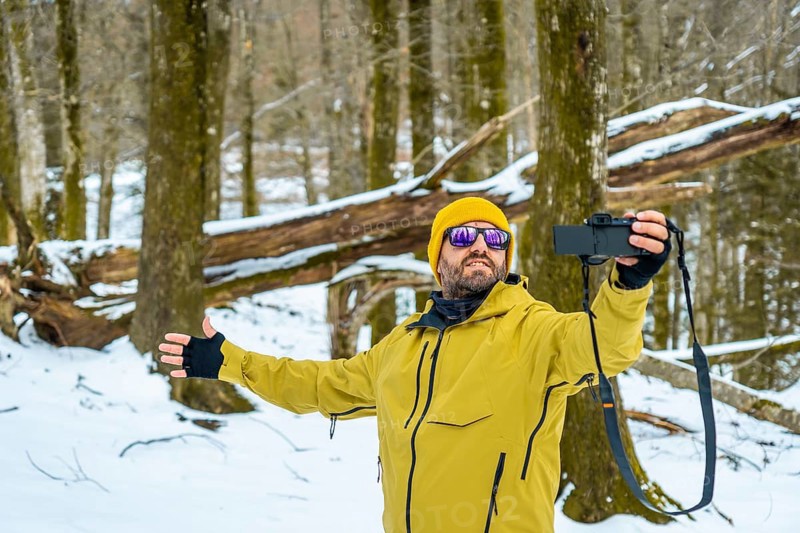 Man wearing a yellow jacket and beanie capturing moments with his camera in a snow covered forest, documenting his winter adventure and sharing his travel experience