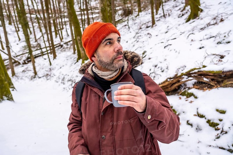 Man wearing an orange beanie and winter jacket, holding a hot mug while contemplating the serene snowy forest, enjoying solitude and warmth during a cold outdoor adventure