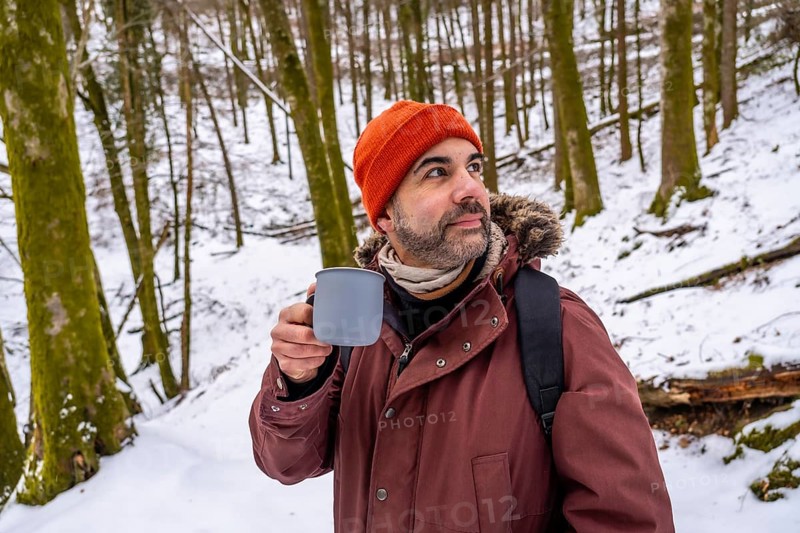 Bearded man in an orange beanie and jacket sips from a mug while standing in a snow covered forest, gazing into the quiet winter landscape and enjoying peaceful solitude