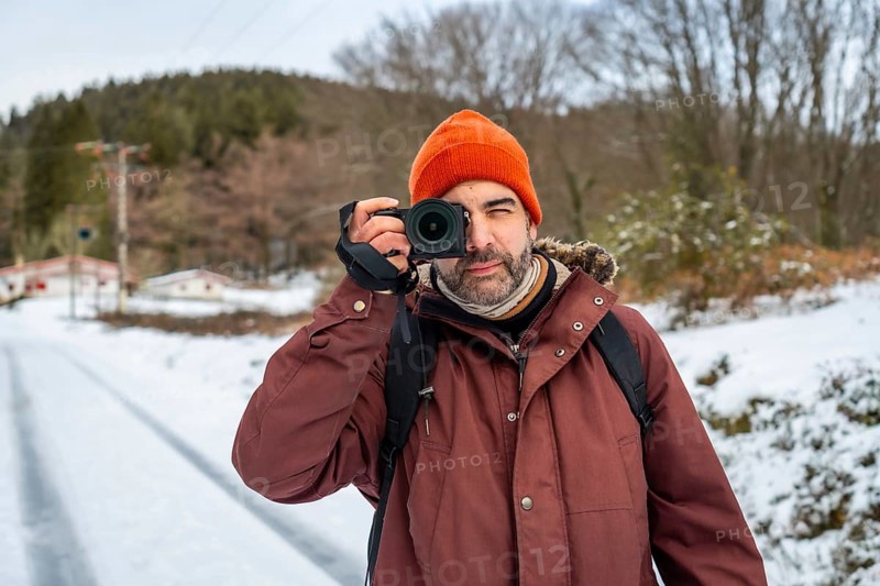 Man in winter clothing and an orange beanie holding a mirrorless camera, capturing nature and winter landscapes on a snowy path during a cold weather adventure