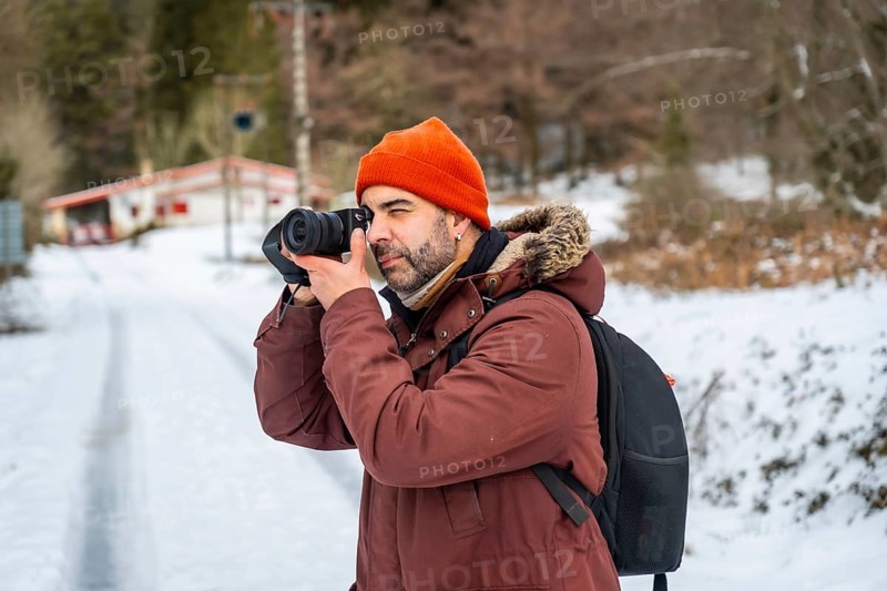 Man wearing an orange beanie and warm winter jacket holding a camera, actively shooting nature and landscapes during a cold, snowy day in the mountains, enjoying outdoor photography