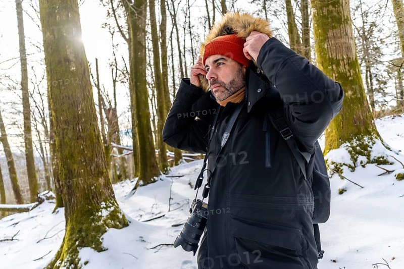 Man photographer adjusting his jacket or coat hood with fur trim while wearing a beanie and scarf outdoors in a snowy forest during winter with trees and sunlight