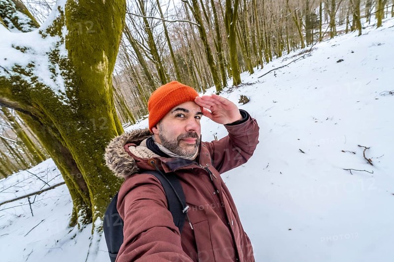 Man posing for a selfie in a snowy forest during winter, looking out with his hand shading his eyes, wearing an orange beanie and a warm jacket for outdoor adventure