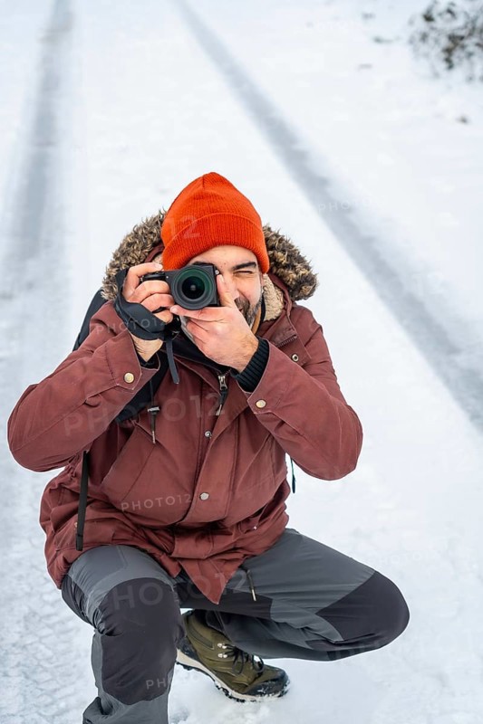 Photographer wearing an orange beanie and parka kneeling in the snow, capturing images with a digital camera during a cold winter day in a natural outdoor environment