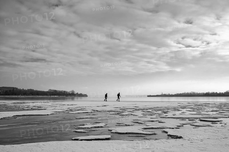 Two people walk across an icy water surface under a cloudy sky, black and white photography, Radolfzell am Lake Constance, Konstanz district, Baden-Württemberg, Germany
