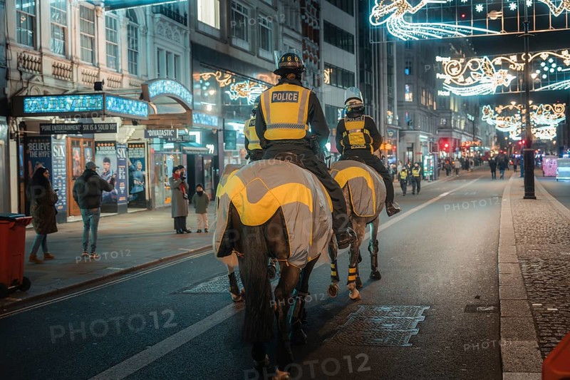 Police horses patrol an illuminated city street at night, surrounded by pedestrians and bright lights, London, England, Great Britain