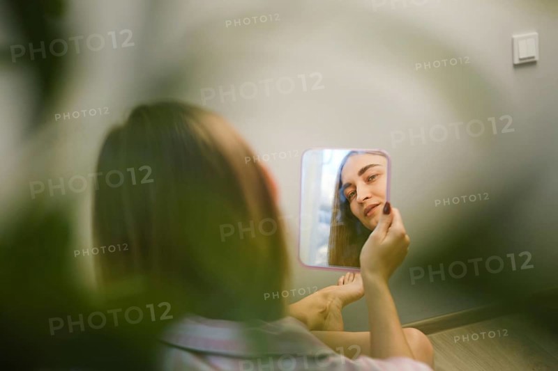 A young woman sits on the floor and looks into a small mirror held in her hand. She is in a bright indoor space surrounded by green plants. Natural light enters through a window