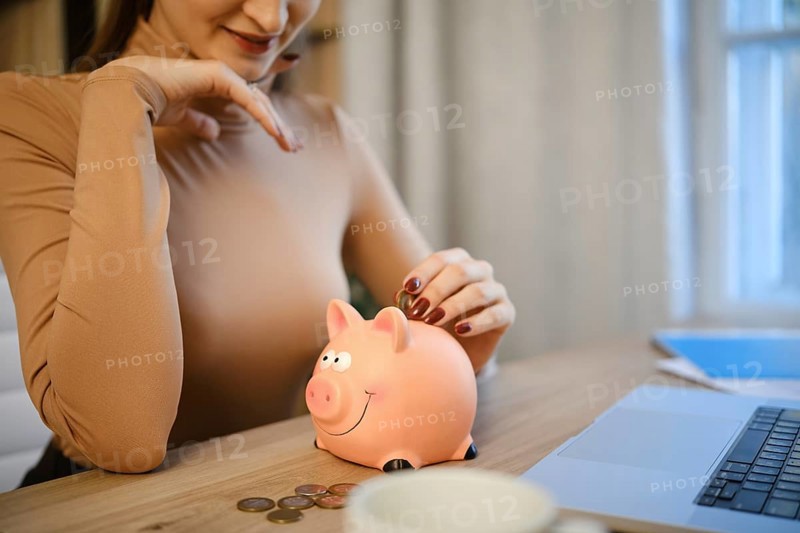 A woman sits at a wooden desk, placing coins into a pink piggy bank. She is focused on saving money while working on her laptop. The room has bright light coming through the windows
