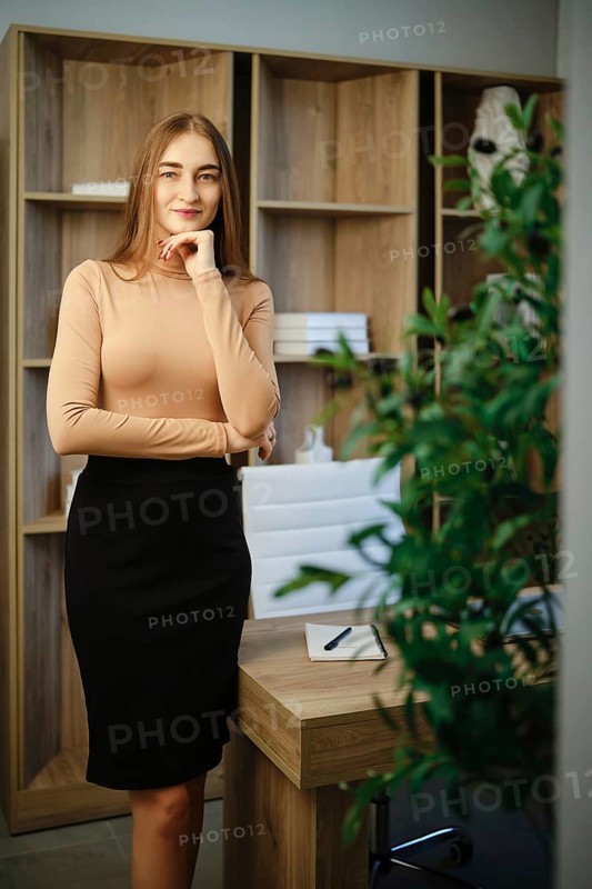 A woman stands in an office with wooden shelves behind her. She has long hair and wears a fitted top with a skirt. A green plant is in the foreground. The setting has a relaxed and inviting vibe