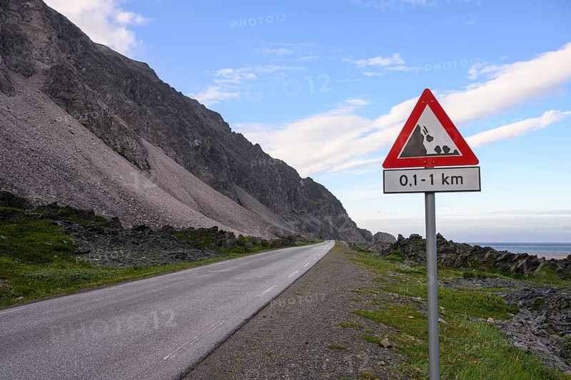 An empty coastal road with a danger sign in front of a mountain range under a blue sky in the north of the Varanger Peninsula, Berlevåg, Finnmark, Norway