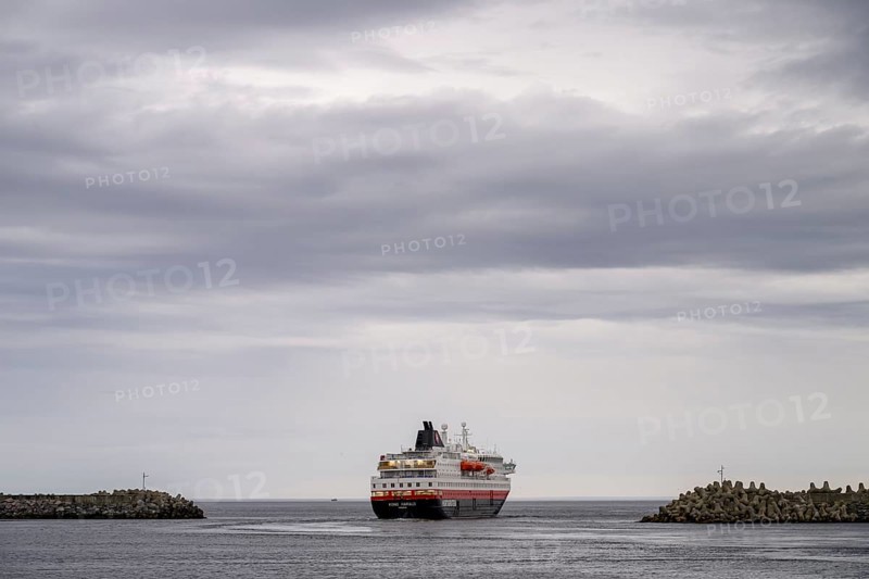 The Hurtigrouten Hurtigruten ship Kong Harald sails from Berlevag harbour, on the horizon with a cloudy sky, Berlevåg, Finnmark, Norway