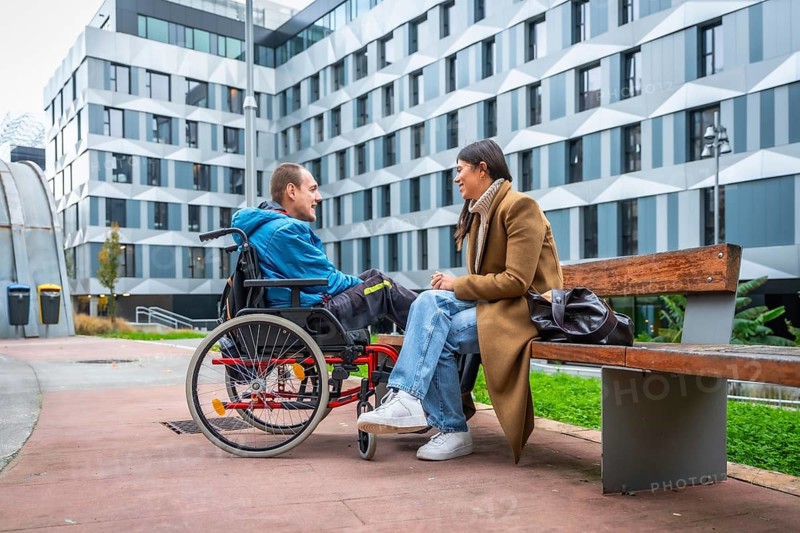 Young man living with a disability sitting in a wheelchair and having a joyful conversation with a woman sitting on a public park bench in an urban setting