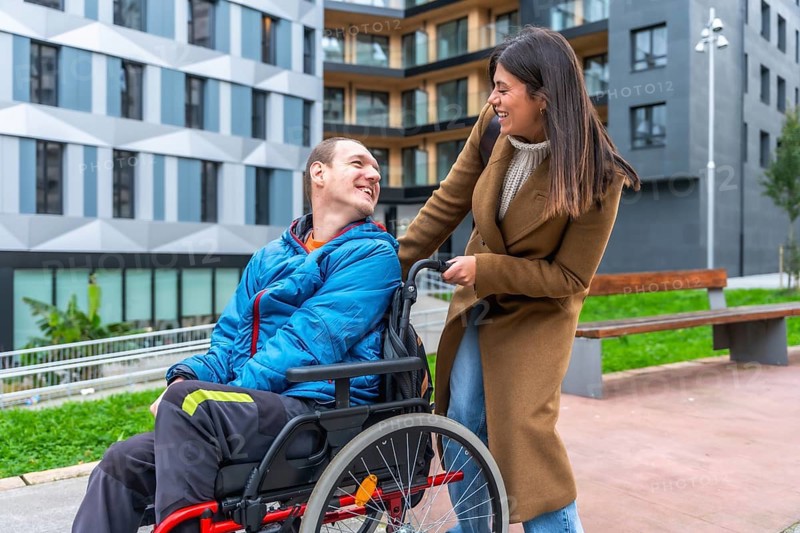 Woman companion assisting a smiling man using a wheelchair, enjoying a happy moment together in an urban park setting, representing support, accessibility, and inclusion