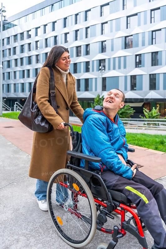 Caregiver pushing a man with a disability in a wheelchair outdoors, both smiling and sharing a happy moment during an urban outing, highlighting inclusion and support in daily life