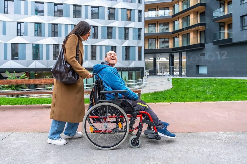 Caregiver pushing a happy adult man with a disability in a wheelchair, enjoying time outdoors together in an urban environment with modern buildings in the background