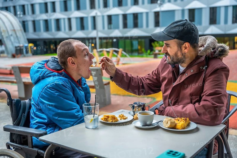 Man in wheelchair being fed by caregiver at an outdoor cafe table, showing compassion, inclusion and everyday support in an urban park setting during a casual meal