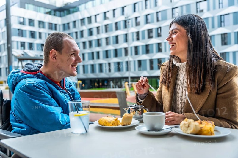 Young man in wheelchair having a joyful conversation and enjoying coffee with a female friend at an outdoor cafe, symbolizing inclusion, friendship, and happy social interaction