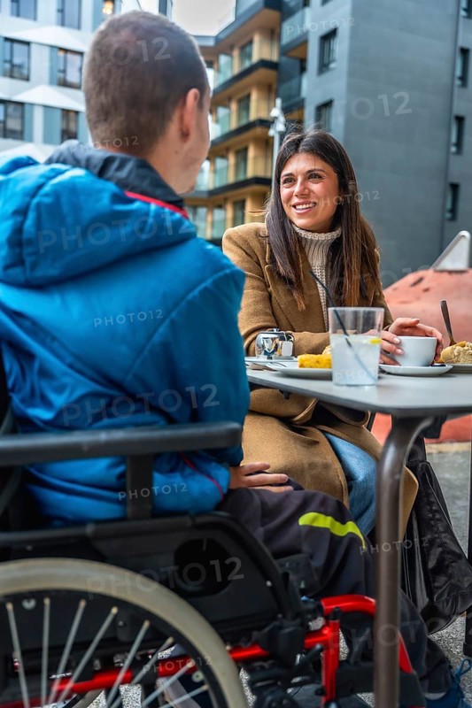 Woman and boy with disabilities enjoying a conversation together while sitting at an outdoor cafe table in an urban setting, sharing an inclusive moment