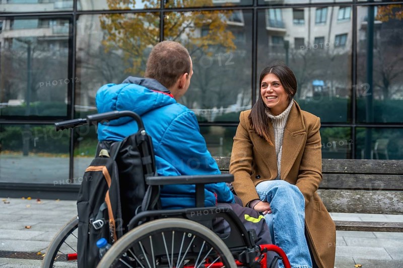 Disabled man seated in a wheelchair engaging in a joyful conversation with a supportive woman on a bench, fostering inclusion and social connection outside