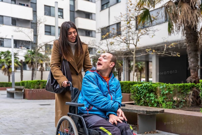 Woman pushing an adult man in a wheelchair, providing support and care during an urban outing, emphasizing challenges and inclusion for people with disabilities