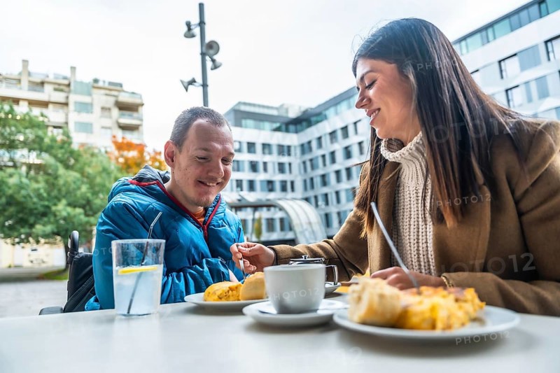 Smiling caregiver offering food to a man in a wheelchair, enjoying a pleasant conversation and meal together at a city cafe, symbolizing support, inclusion, and everyday social life