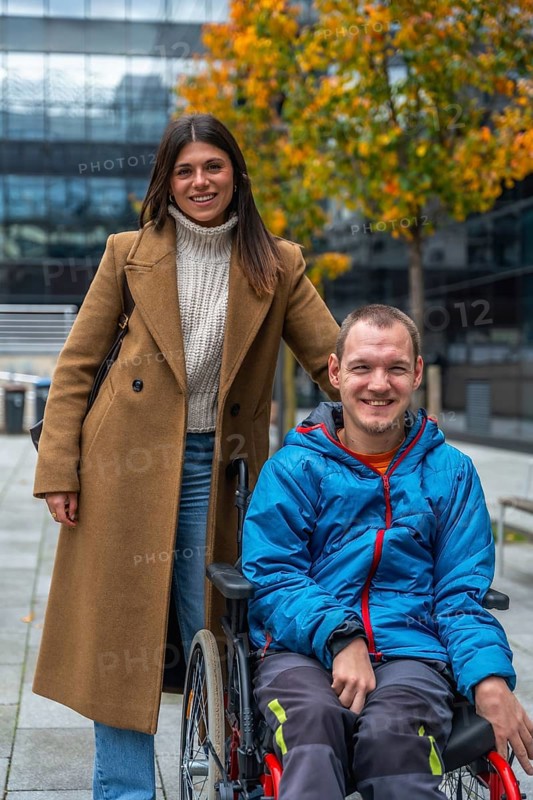 Young woman pushes her smiling partner in a wheelchair along an accessible urban pathway amid autumn foliage, sharing support, independence and joyful outdoor moments
