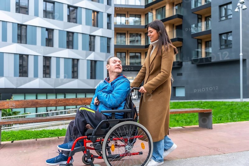 Young woman smiling as she pushes a man in a wheelchair along an accessible city street, showcasing inclusion, support, mobility and independent urban life together