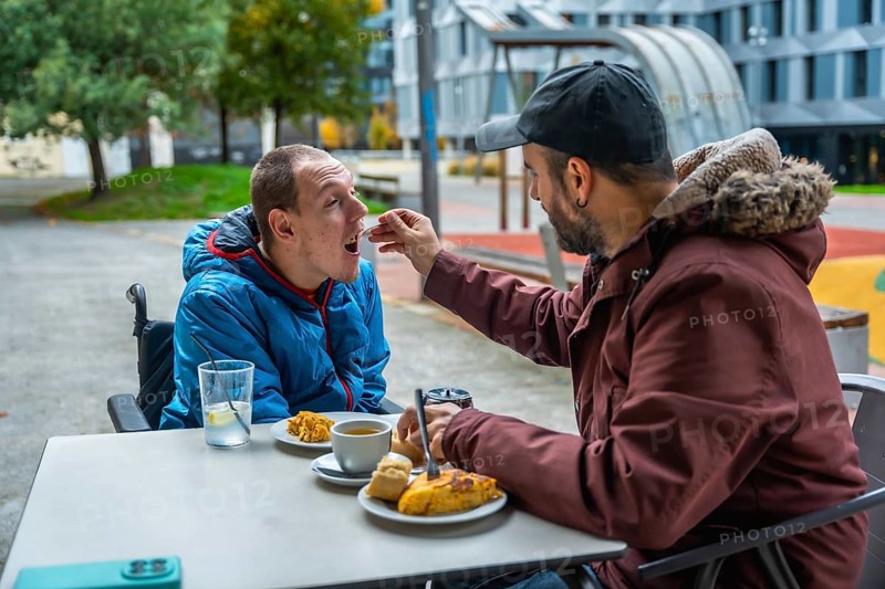 Man tenderly feeding a young boy in a wheelchair at an outdoor cafe, representing care, support, and inclusion for people with disabilities in public spaces