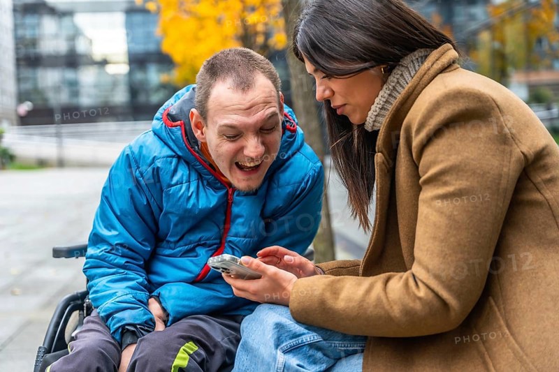 Disabled man in a wheelchair smiling with a female caretaker enjoying a fun moment together while looking at a smartphone screen outside during autumn