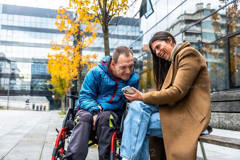 Man in a wheelchair and woman sitting on a bench, sharing a laugh and looking at a mobile phone screen outdoors in an urban setting with autumn foliage