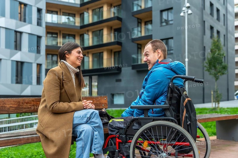 Happy young woman sitting on a bench, engaging in conversation and laughing with a smiling man using a wheelchair, showing friendship and inclusion in an urban setting