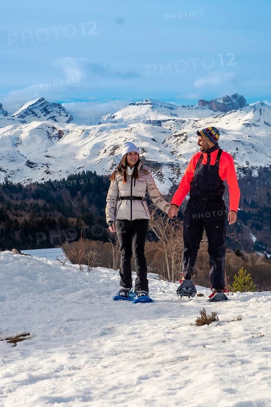 Young couple hand in hand snowshoeing on a sunny mountain trail, surrounded by vast snow capped peaks and blue sky, enjoying winter adventure and fresh alpine air