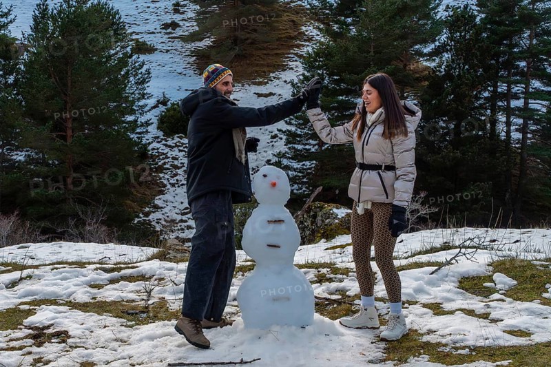 Young couple enjoying winter vacation, high fiving, and smiling while building a snowman on a snowy hillside with pine trees and mountains in the background