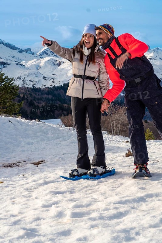 Smiling couple snowshoeing across snowy mountain slopes, pointing toward a breathtaking winter panorama under blue skies, enjoying active outdoor adventure and fresh alpine air