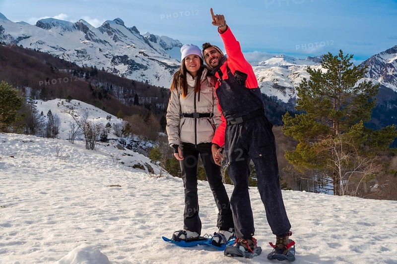 Young couple enjoying outdoor winter adventure, snowshoeing in a snowy mountain landscape under blue skies, with the man pointing excitedly towards something in the distance