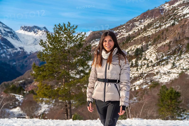 Young woman smiling and walking through snowy mountain landscape, wearing a warm jacket and scarf while enjoying fresh air, sunshine and scenic winter hiking adventure