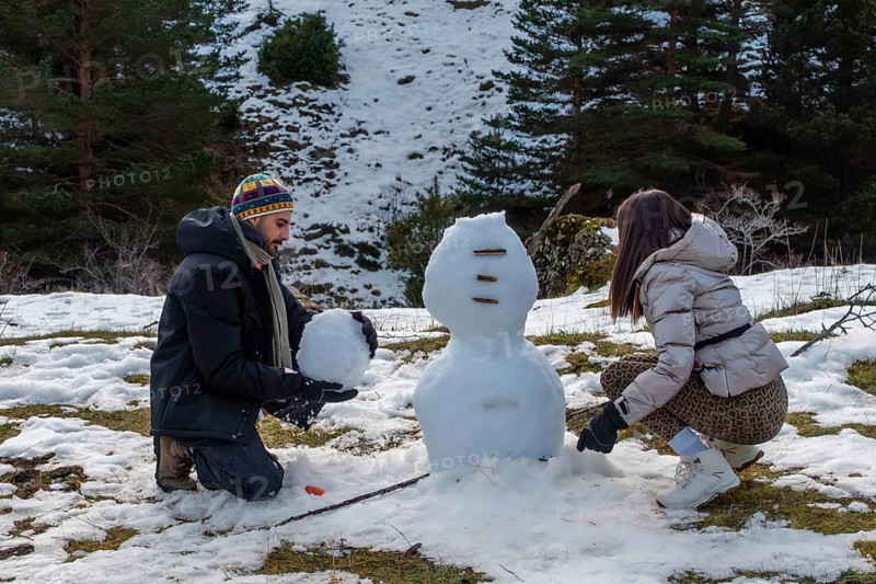 Young couple building a snowman on a sunny mountain slope, laughing and bonding in warm winter clothes amid snowy forest scenery, enjoying playful outdoor holiday recreation