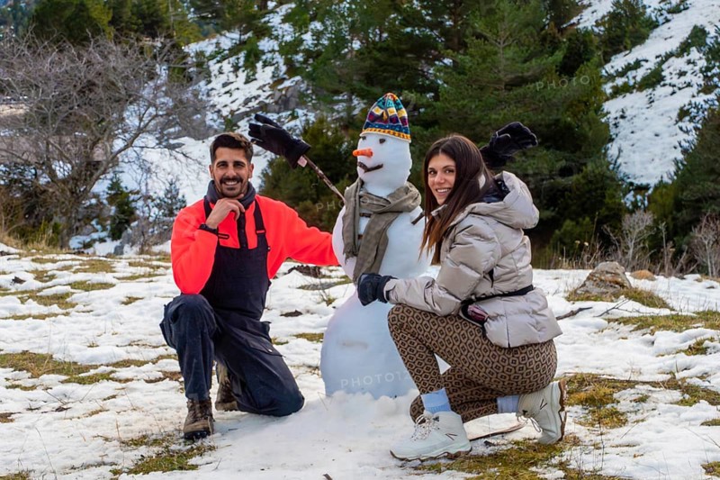 Smiling couple crouches beside a snowman in a snowy mountain forest, enjoying playful winter holiday moments together with scarves, hats and warm laughter outdoors