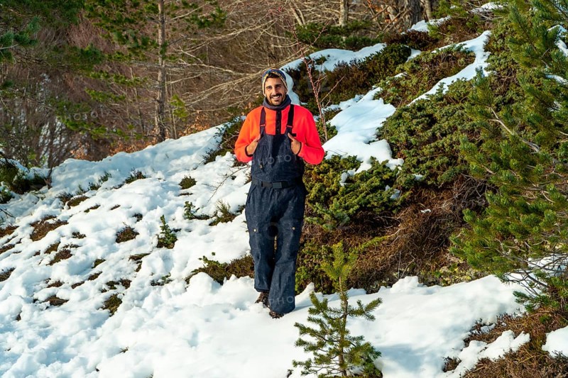 Man in an orange sweater and dark bib pants smiling, enjoying winter nature while walking through a snowy landscape with green bushes and pine trees under sunlight