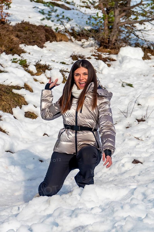 Woman expressing joy and playfulness while preparing to throw a snowball outdoors, dressed in warm metallic winter clothing against a snowy mountain backdrop