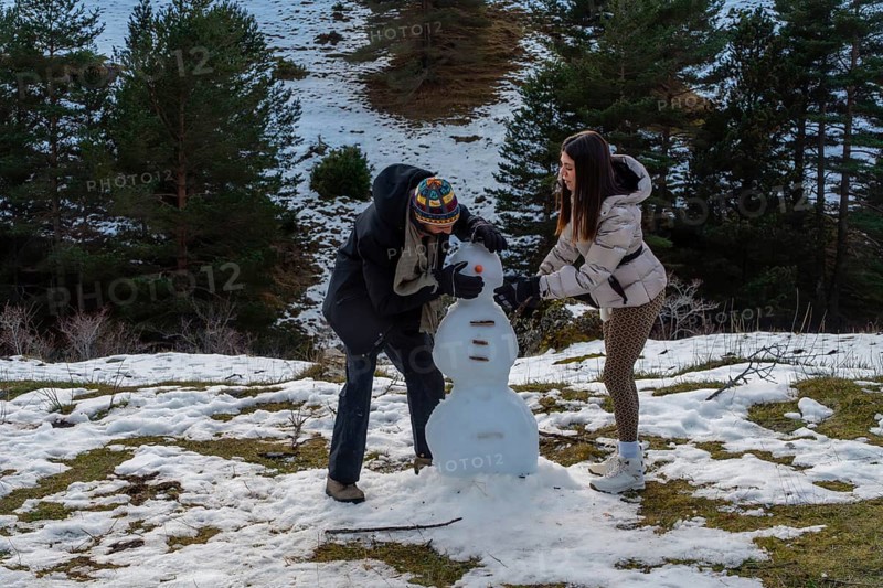 Young couple building a snowman in a snowy mountain pine forest, laughing and bonding outdoors on a winter getaway, enjoying playful recreation and fresh air together
