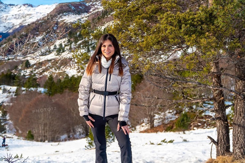 Woman posing in a warm jacket and ski pants, smiling at the camera, exploring a snowy mountain landscape with pine trees and clear sky in the background, embracing winter adventure