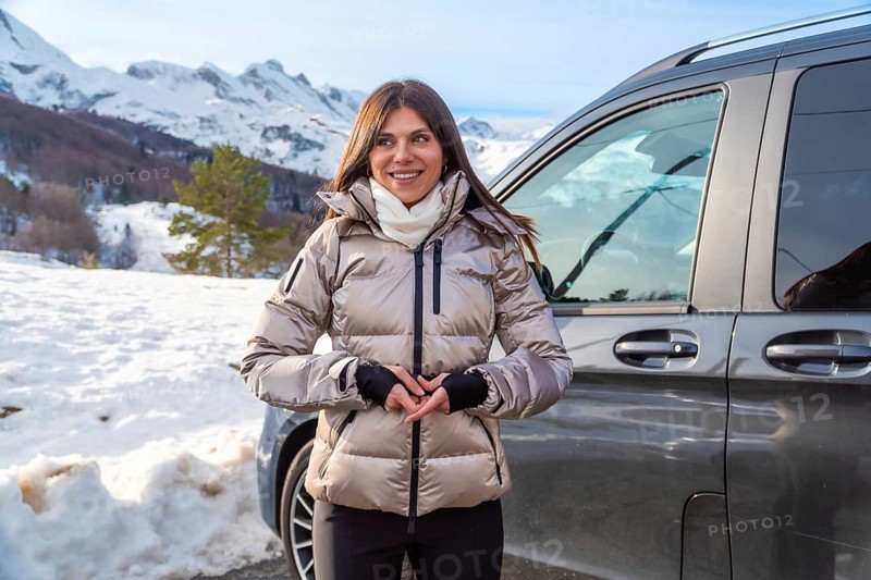 Woman standing by a vehicle against snowy mountain peaks, wearing a warm jacket and scarf, smiling and enjoying a scenic winter road trip and outdoor adventure