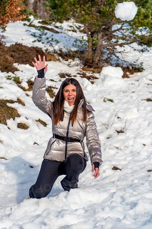 Young woman kneeling in snow with a playful expression, throwing a snowball against a backdrop of snow covered terrain and trees, embodying winter fun and outdoor activity