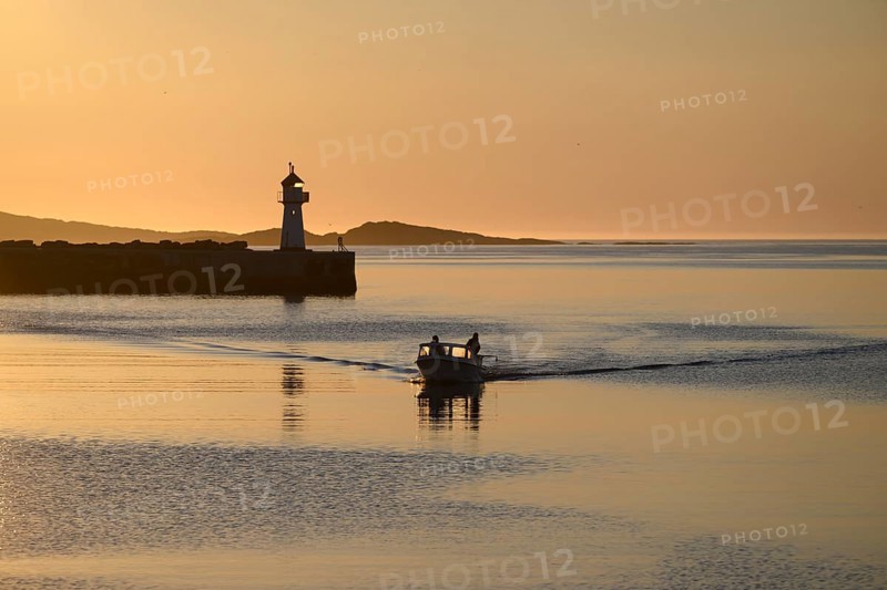 A small motorboat sails through the harbor entrance with lighthouse and midnight sun, Vardø, Finnmark, Norway