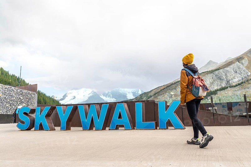 Female tourist walking near the skywalk sign with the athabasca glacier and mountains in the background at the columbia icefield in jasper national park, alberta, canada